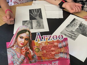 Hände liegen auf einem Tisch mit Henna-Verpackung, Henna-Stiften und ausgedruckten Vorlagen in Schwarz-Weiß.