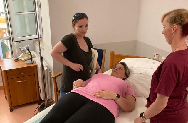 Frau liegt auf dem Rücken in einem Pflegebett. Zwei Frauen stehen zu beden Seiten am Bett.