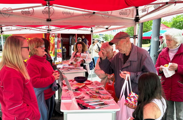 AWO-Info Stand unter rotem Pavillon. Links vom Tisch Mitarbeiterinnen der AWO. Rechts vom Tisch ältere Menschen die sich informieren. Auf dem Tisch viel Info-Material.