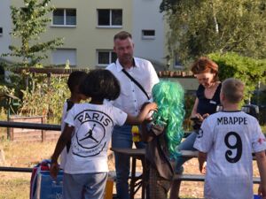 Draußen. Vier Kinder und zwei Erwachsene. Mann in weißem Hemd und Jeans steht an einem Tisch. Drei schwarze Kinder, eines trägt eine Perücke lange grüne Haare. Ein weißer Junge mit Fußball Trikot kommt dazu.