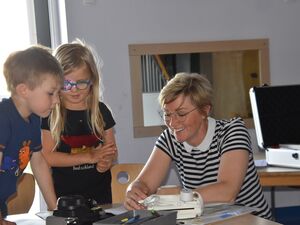 Junge und Mädchen am Tisch. Sie gucken neugierig. Eine Frau sitzt am Tisch und lächelt. Die hat etwas vor sich. Die Kinder sehen zu. Das Mädchen trägt ein schwarzes T-Shirt mit Deutschlandflagge, der Junge mit die Maus und dem kleinen Elefant.