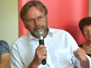 Nahaufnahme. Mann mit Mikrofon in der rechten Hand. Mann hat braune Haare, Vollbart und eine Brille auf. Er trägt ein weißes Hemd mit Kragen.