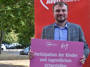 Portrait: Mann mit braunen kurzen Haaren, freundlichem Lächeln und Drei-Tage-Bart. Er trägt ein blaues Hemd mit Kragen darüber ein graues Jackett. Er hält ein pinkes Schild vor sich. Text: Partizipation der Kinder und Jugendlichen sicherstellen.