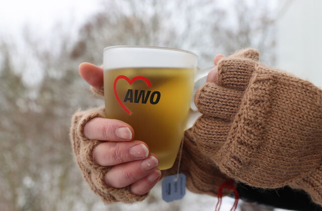 Teetasse mit AWO-Logo. Tasse wird mit zwei Händen in fingerlosen braunen Handschuhen gehalten.