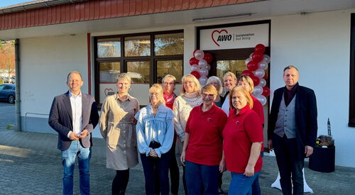 11 Personen, zwei Männer links und rechts außen stehen vor einem weißen Gebäude. Der Eingang ist mit roten und weisen Luftballons geschmückt.