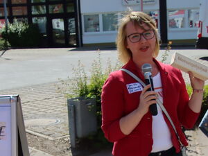Im Freien. Frau mit Mikrofon in der Hand. Sie ist blond, trägt eine Brille, ein weißes T-Shit und darüber einen roten Blazer.