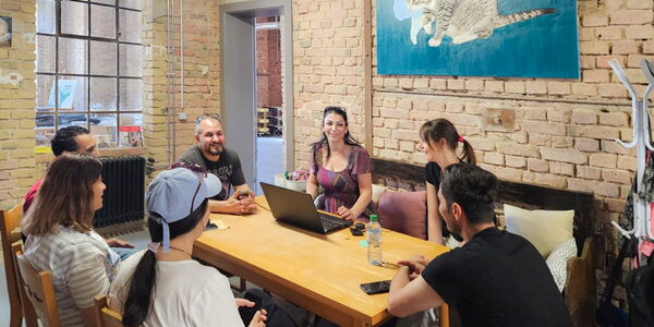 7 Menschen sitzen um einen Tisch. Eine Frau auf einer Bank sitzend, hat vor sich einen Laptop. An der Wand ist ein großes blaues Bild mit einer Katze in der Mitte.
