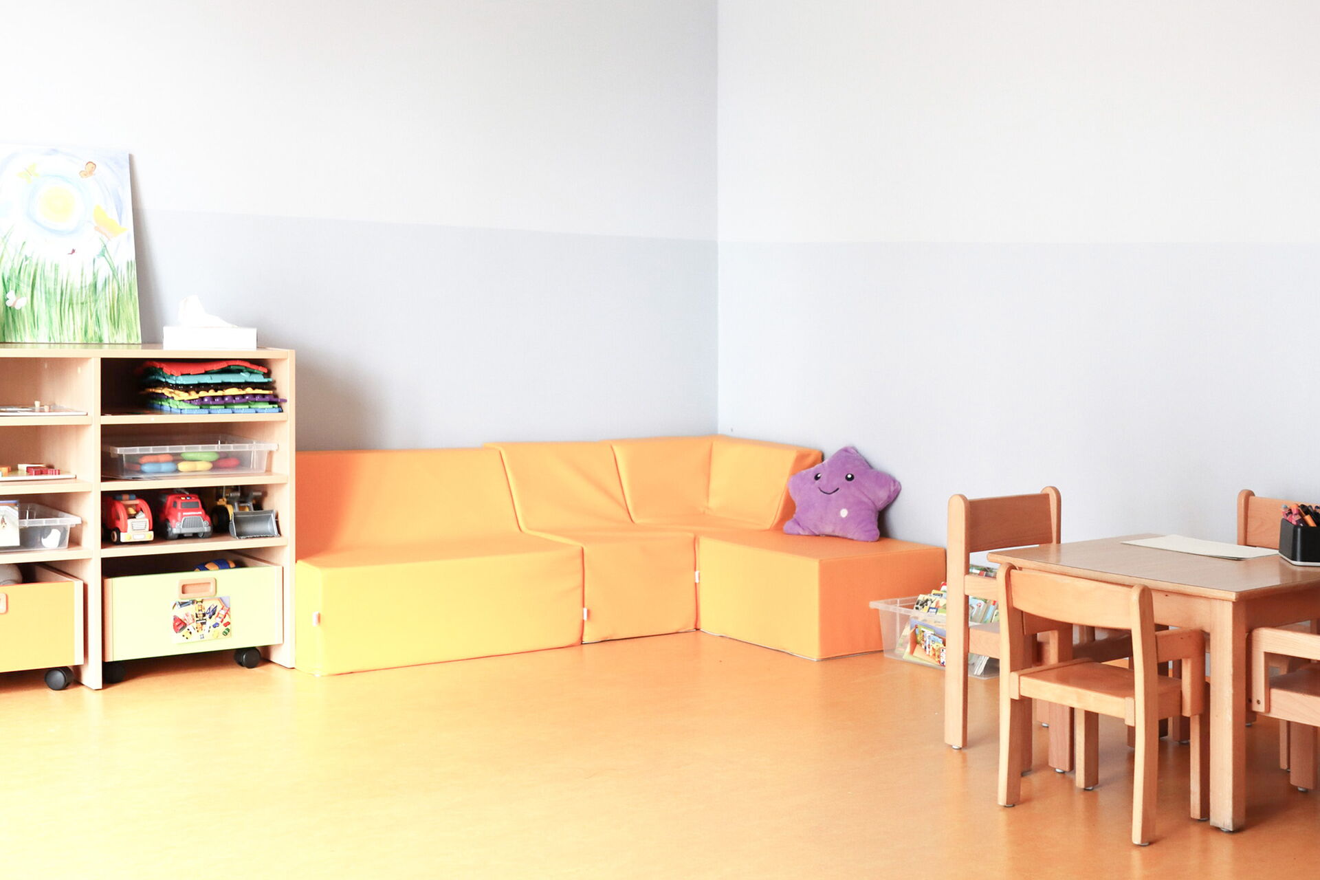 Freizeitraum. Freundlich, hell. An der Wand rechts ein kleiner Tisch mit 4 Stühlen. In der Eche eine gemütliche orangene Sitzecke mit einem lila Plüsch-Stern darauf. Daneben ein kleines Regal.