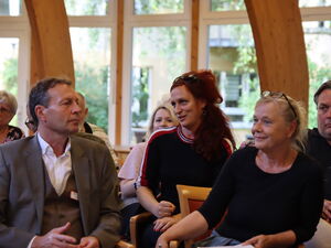 Menschen sitzen auf Stühlen, In der ersten Reihe ein Mann im Anzug und eine blonde Frau mit Brille auf den Kopf geschoben.