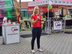 Blonde Frau mit Mikrofon in der rechten Hand und eine Kamera in der linken Hand. Sie trägt ein rotes T-shirt und lange blonge glatte Haare ohne Pony. Dahinter im AWO Pavillon der Techniker am Pult.