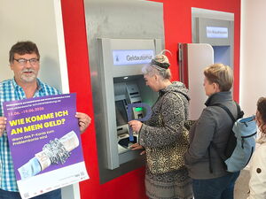 Menschen-Schlange vor Geldautomat. Einige Frauen halten ein großes P In die Kamera. Ein Mann steht links außen und hält ein lila Plakat "Wie komme ich an mein Geld?"