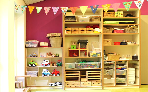 Heller Raum, großes Regal steht an der Wand. Körbe und Aufbewahrungskisten im Regal. links daneben in der Ecke ein kleines Regal mit Spielzeug, Autos, Bagger