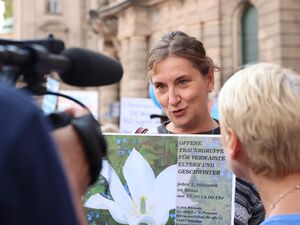 Eine TV-Kamera mit Mikrofon ist auf eine frau gerichtet. Die Frau hat braune Haare. Sie hält ein Schild der offenen Trauergruppe für verwaiste Eltern und Geschwister vor sich.