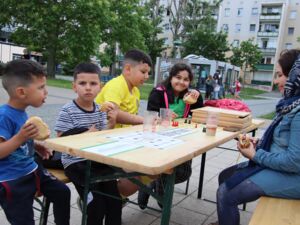 Biertischgarnitur an der 3 Jungen und ein Mädchen sitzen. Gegenüber der Kinder sitzt eine in Jeans Kleidung und mit Kopftuch. Sie essen Burger und spielen ein Brettspiel.