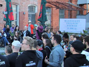 Viele Menschen vor einem rotenn Backsteingebäude. In der Mitte der Menschen sind Musikerinnen mit Gitarren. Viele Leute tragen Kleidung mit Aufdruck vom Potsdamer Kneipenchor.