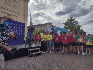 Mannn auf kleiner Bühne spricht in ein Mikro. Gebärdendolmetscherin steht auf der Bühne. Menschen stehen daneben und einer im roten T-Shirt und schwarzer kurzer Hose hält eine brannende Fackel in die Höhe.