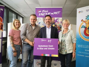 Gruppenbild vor dem lila Roll up Banner vom Büro Kinder(ar)mut. 2 Männer in der Mitte, 2 Frauen. Fischer hält lila Schild mit Text: Kostenloses Essen in Kitas und Schulen.