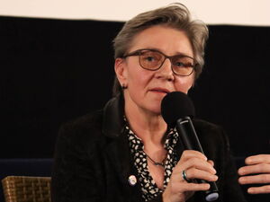 Frau sitzt auf einem Stuhl - Podiumsdiskussion . Sie hält Mikro in der Hand. Sie hat kurze Haare, Brille, und trägt dunkle Kleidung.