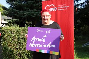 Portrait: Draußen. Frau mit roten kurzen Haaaren, roter Brille, roten Lippen und roten Fingernägeln hält ein lila Schild in den Händen. Sie steht vor einem roten AWO RollUp Banner. Auf dem Schild steht: Armut überwinden.
