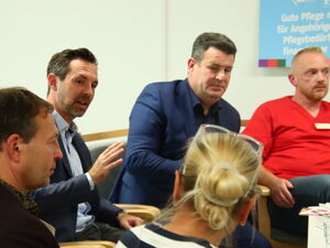 Stuhlkreis von nahem, Zwei Männer im Anzug und einer mit rotem AWO Shirt. An der Wand hellblaues Schild Forderung: Gute Pflege muss für Angehörige und Pflegebedürftige finanzierbar sein.