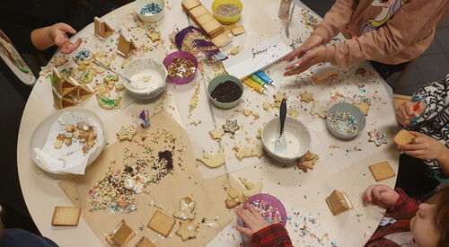 Runder Tisch von Oben. Kinderhände dekorieren Kekse / Plätzchen. Auf dem Tisch sind Butterkekse, Zuckerguss, bunten Zuckerstreuseln, Plätzchen. Alles ist auf dem Tisch verstreut.