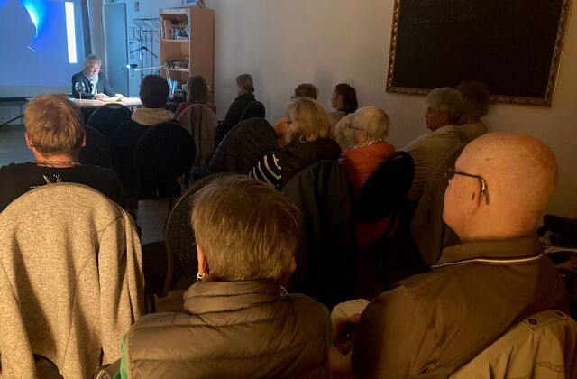 Ca. 15 Menschen sitzen in einem Raum. Vorne ist ein Autor der aus seinem Buch liest. Der Tisch an dem er sitzt ist beleuchtet. Der restliche Raum ist eher dunkel. Wenige Lichtquellen.
