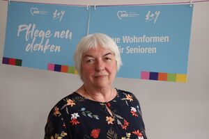 Portrait. Ältere Frau mit weißen kurzen Haaren, eine dunklen Bluse mit farbenfrohen Blüten und Blättern. Hinter ihr hängen zwei Schilder: Pflege neu denken | Neue Wohnformen für Senioren.