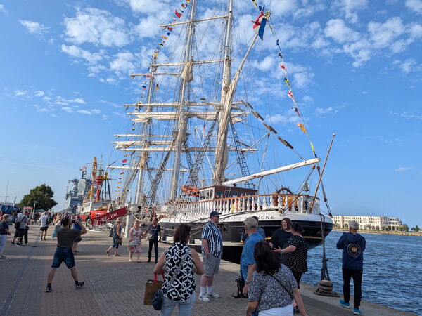 Großes Segelschiff mit hohen Masten liegt am Kai, mehrere Menschen stehen und gehen auf der Uferpromenade.