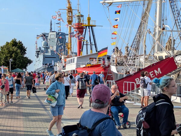 Menschenmenge auf einer Uferpromenade vor einem großen Segelschiff mit deutscher Flagge, rechts rote Gangway mit Schriftzug.