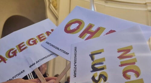 Eine Hand hält drei kleine Protestschilder in einer Hand. Auf den Schildern steht: Nicht lustig, Oha und Dagegen