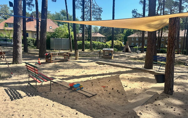 Sandkasten überdacht mit gelben Sonnensegel das an umstehenden Bäumen befestigt ist.