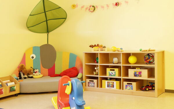 Kleiner bunter Schaukel-Elefant. Ein Elefanten Sitzecke und ein Regal mit Spielzeug steht an einer gelben Wand.