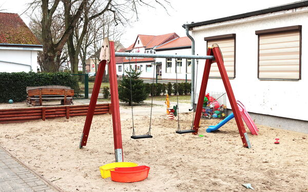 Großer Sandkasten mit zwei Schaukeln, Rutsche für Kleinkinder. Dahinter Holzbänke und eine Hecke.