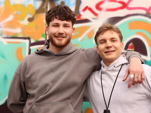Zwei junge Männer, vor einer Graffiti-Wand. Beide lächeln in die Kamera.