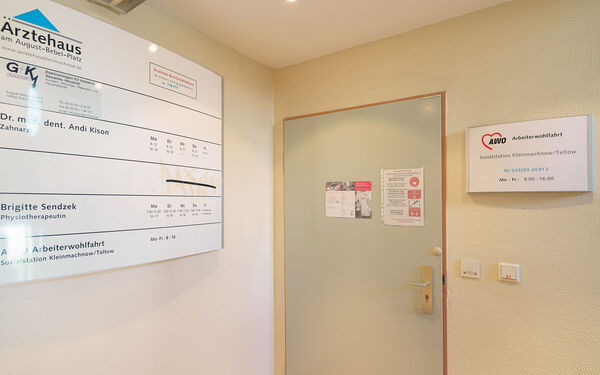 Flur. Geschlossene Tür zur Sozialstation. Links an der Wand ein großes Schild vom Ärztehaus worin sich die Sozialstation befindet.