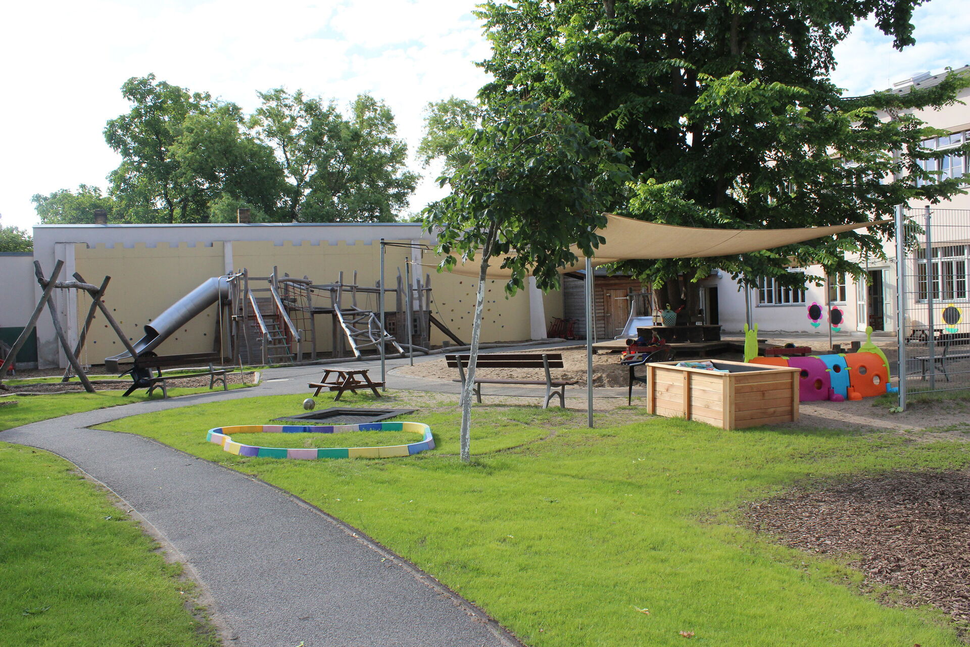 Draußen, großer Spielplatz mit Schaukeln, Klettergerüst mit Rutsche, Sandkasten. Ein großer Baum und ein Sonnensegel spenden Schatten.