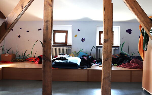 Dachgeschoss. Kleine Fenster. Großer Spielraum der von Holzbalken gestützt wird. Auf einer erhöhten Holzfläche liegen Decken, Kissen und kleidung chaotisch durcheinander.