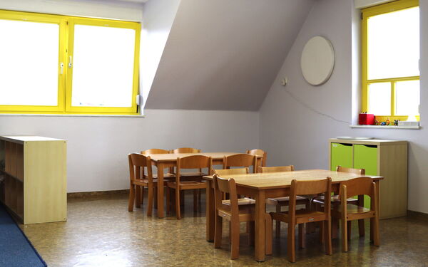 Heller Raum mit zwei kleinen Tischen mit jeweils 6 kleinen Stühlen. Ein kleines Schränkchen steht an der Wand unter einem der Fenster mit gelben Fensterrahme.