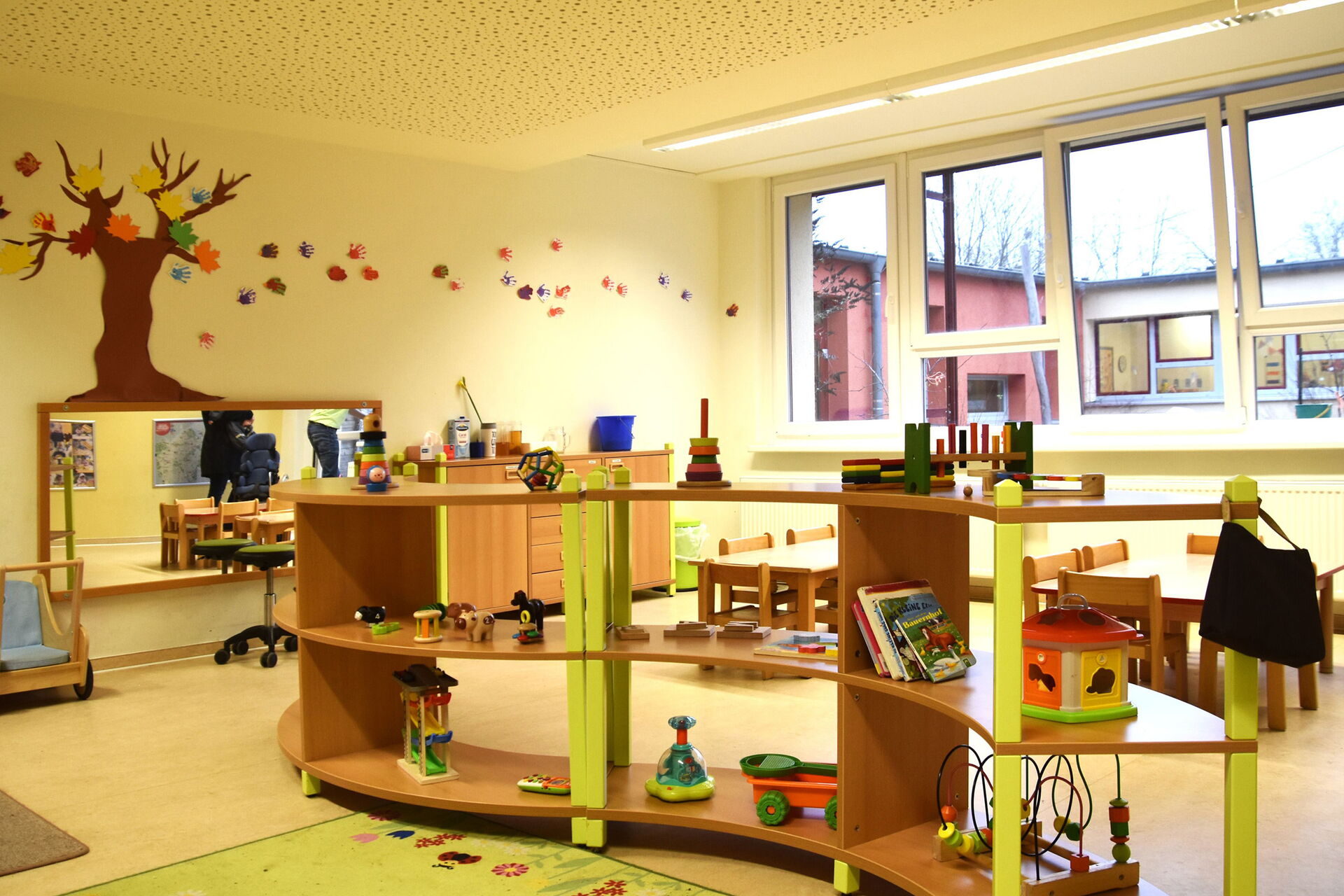 Spielzimmer. Geschwungenes Regal mit Spielsachen für Kleinkinder. An der Wand hinten ein rechteckiger Spiegel. Darüber an der Wand ein kahler Baum mit bunten Herbstblättern aus Papier.