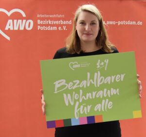 Portrait: Frau, lange blonde Haare, schwarze leidung, freundliches Gesicht. Sie steh vor einem roten AWO Hintergrund. Sie hält ein grünes Schild mit einer AWO Forderung vor sich:    Bezahlbarer Wohnraum für alle