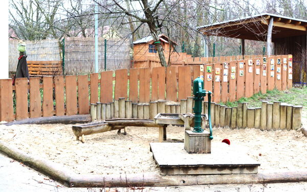 Draußen, kahle Bäume. Ein Wasserspielplatz. Sandkasten, eine Wasserpumpe mit Wasserbecken und zwei Wasserrinnen.