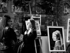 Schwarz-Weiß Foto: Zwei Frauen stehen in Atelier und unterhalten sich. Sie sehen sich an. Eine hält ein Glas in der Hand. Beide haben lange glatte Haare und tragen dunkle Kleidung.