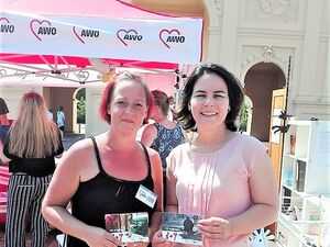 Draußen, vor dem Brandeburger Tor in Potsdam. Zwei Frauen stehen nebeneinander, sie halten Karten in die Kamera und lächeln. Sie sind sommerlich gekleidet. Hinter ihnen ist ein AWO Pavillon.