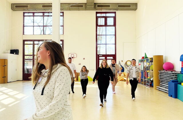 Eine Turnhalle  mit hellen Wänden. Junge Menschen schreiten durch die Halle. Kommen auf die Kamera zu.