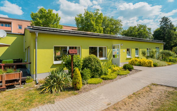 Grünes flaches Gebäude, davor Pflanzen und Büche, die Eingangstür steht offen.