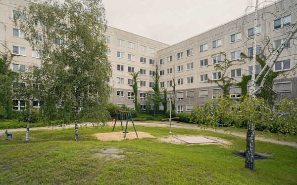 Draußen. Spielplatz, Schaukel, Wippe, Wiese, ein paar Bäume. Grauer Gebäudekomplex an einigen Stellen mit Efeu bewachsen.
