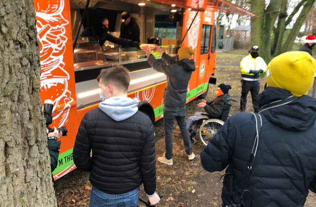 Orangener Food Truck. Menschen in Winterjacken, Mützen und Masken stehen davor. Einer nimmt gerade Essen aus dem Truck entgegen. Ein Rollstuhlfahrer steht neben der Person und streckt die Hände aus um das Essen entgegenzunehmen.