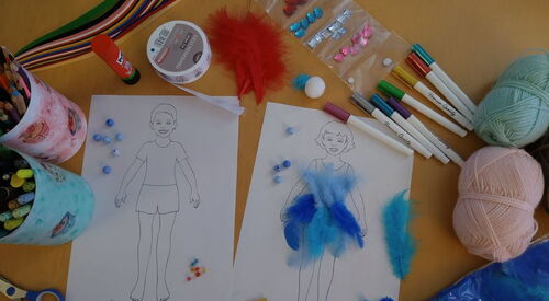Tisch von oben mit Mal- und Bastelutensilien. Zwei Bilder nebeneinander, Junge und Mädchen. Auf dem Bild des Mädchens sind blaue Federn geklebt. Stifte, Schere, Wollknäuel, blaue Perlen, Prittstift