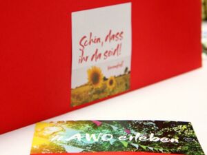 Karte an roten Hintergrund geklebt. Auf der Karte Sonnenblumen und der Text: Schön, dass ihr da seid! Davor auf einem weißen Tisch liegt die Einladung zum Vereinsfest des AWO Bezirksverband Potsdam e.V.