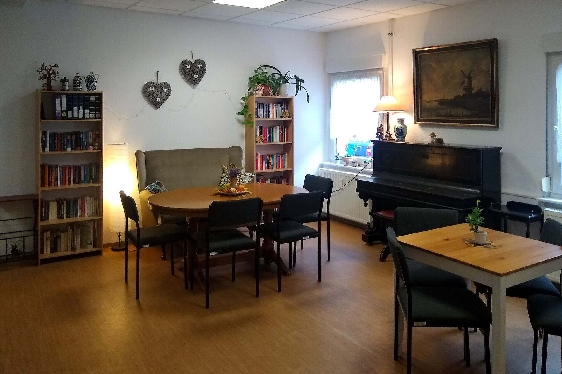 Dunkler Raum, mit Tischen, Stühlen und Sofa. 2 Bücherregale stehen an der Wand.